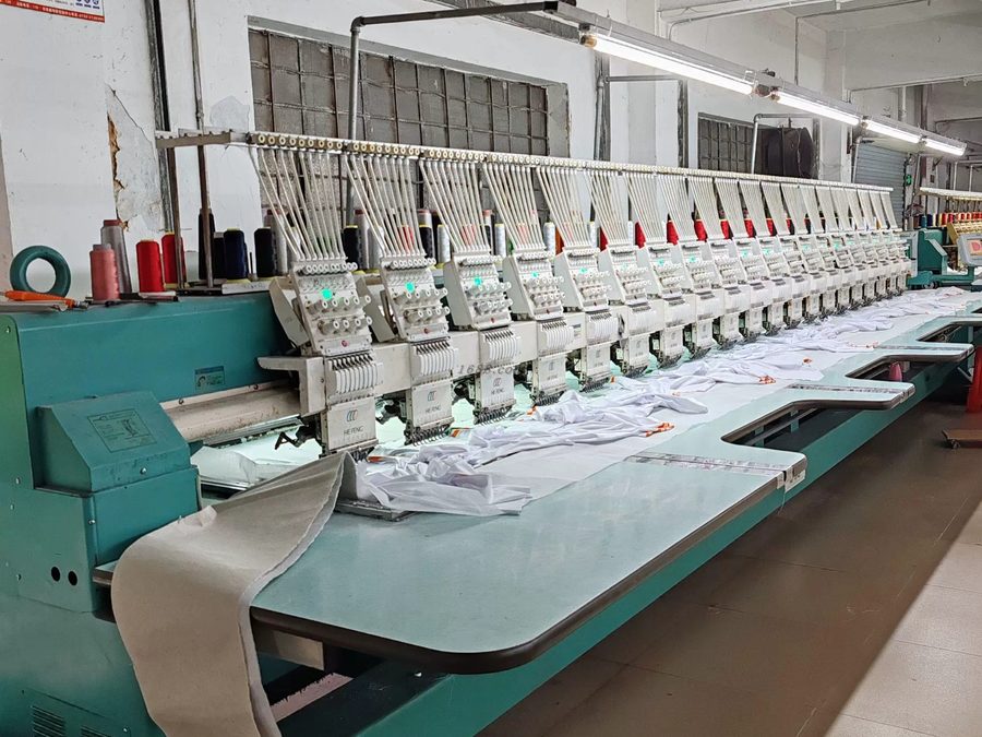 Row of multi-head embroidery machine heads stitching white polo shirts with red and white thread