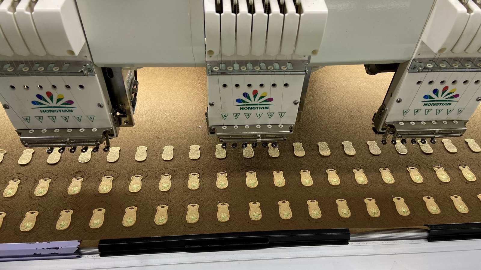 Three embroidery machine heads stitching rows of small cream patch shapes on brown felt