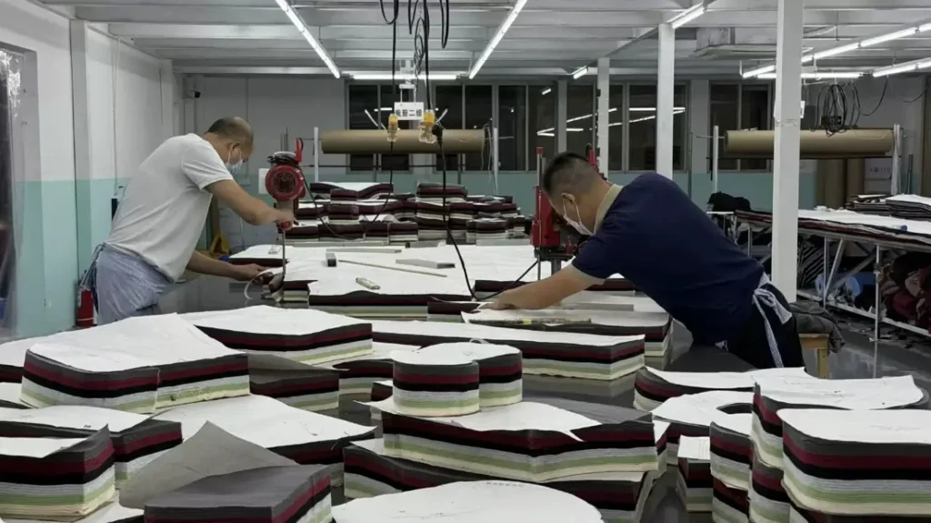 Two workers hand-cutting stacked fabric plies with a round-knife cutter on the factory floor