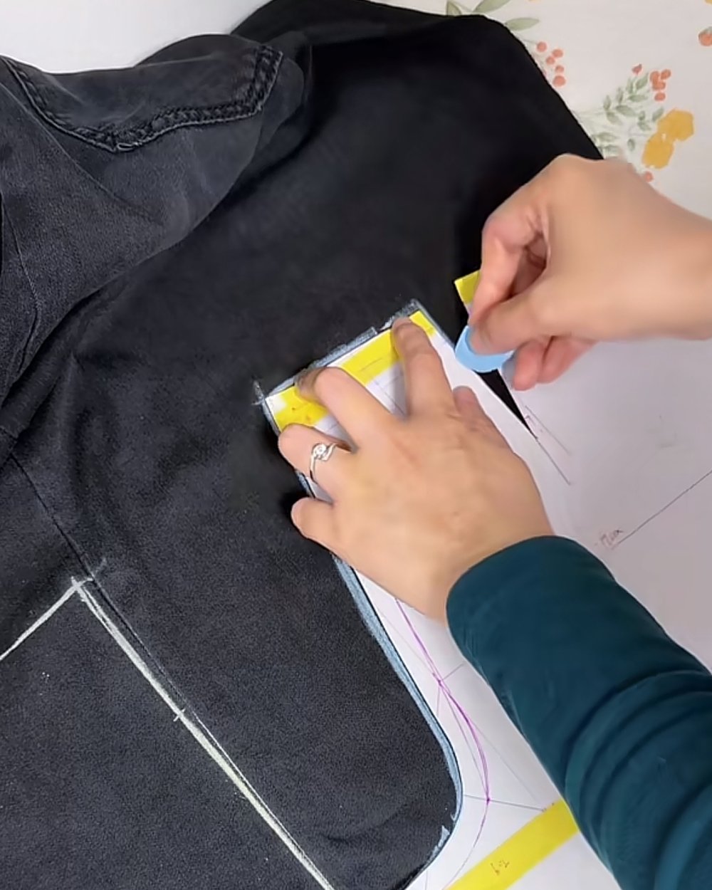 Hands holding a paper template and blue chalk against a washed black denim jacket front, chalk outline drawn below