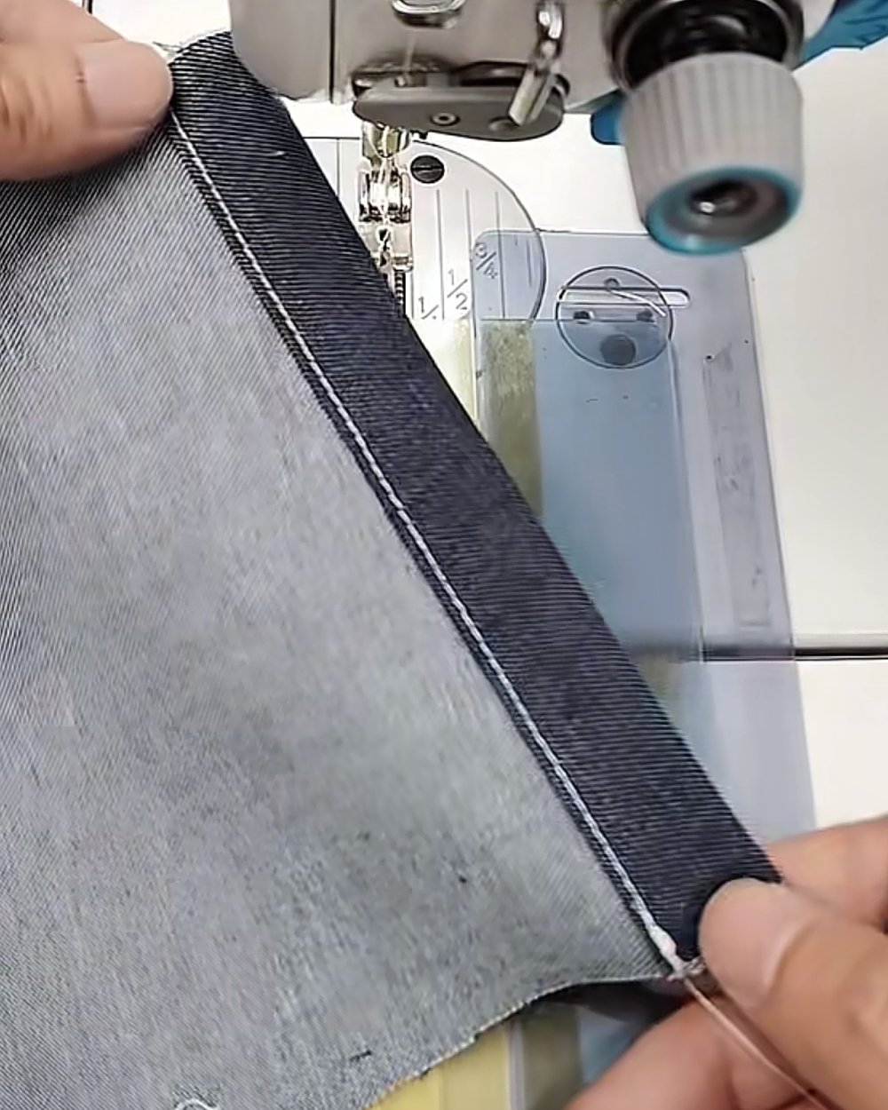 Hands guiding a folded indigo twill hem under the presser foot of a sewing machine, white topstitch visible