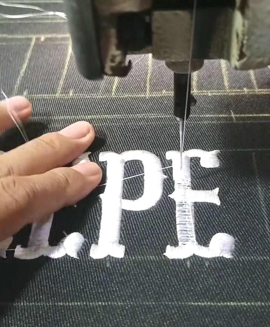 Embroidery machine needle stitching white letters onto dark woven fabric, close-up of the stitching process on our in-house embroidery line
