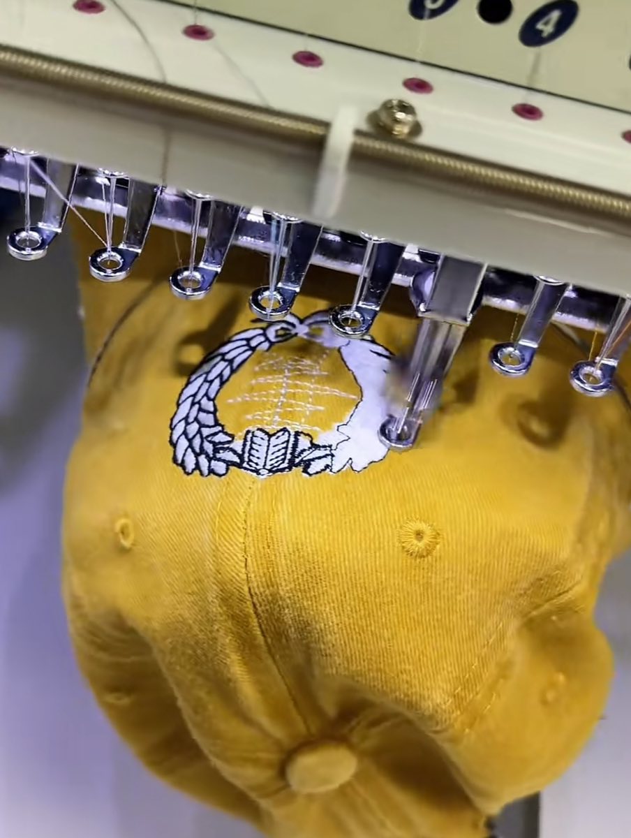 Embroidery machine stitching a white crest motif onto a mustard yellow baseball cap on our in-house embroidery line